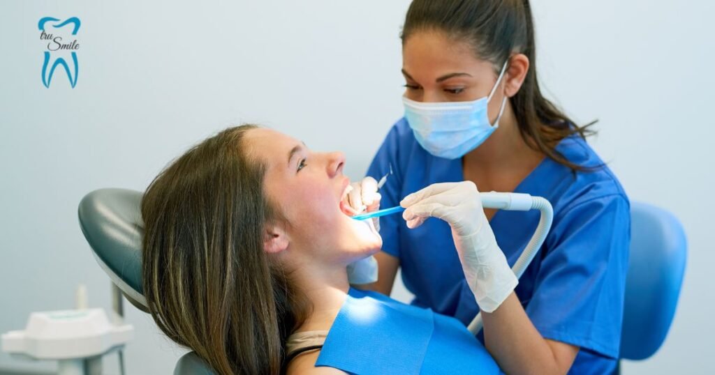 Dental professional in blue scrubs performing a procedure on a patient in a dental chair. The 'tru Smile' logo is visible on the wall.